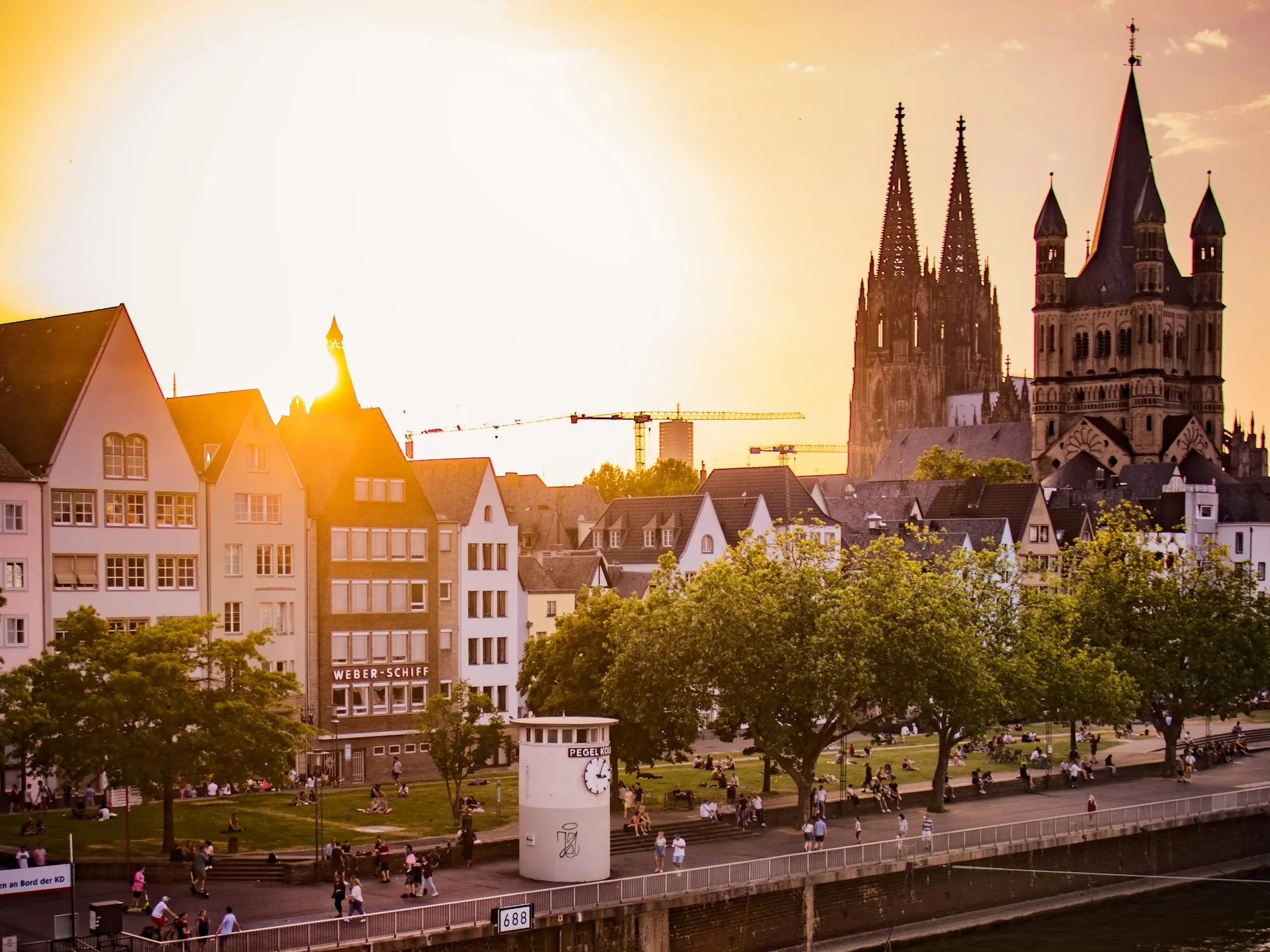 Menschen in Köln spazieren am Rhein entlang oder sitzen in kleinen Gruppen auf einer Rasenfläche, im Hintergrund ist der Kölner Dom zu sehen