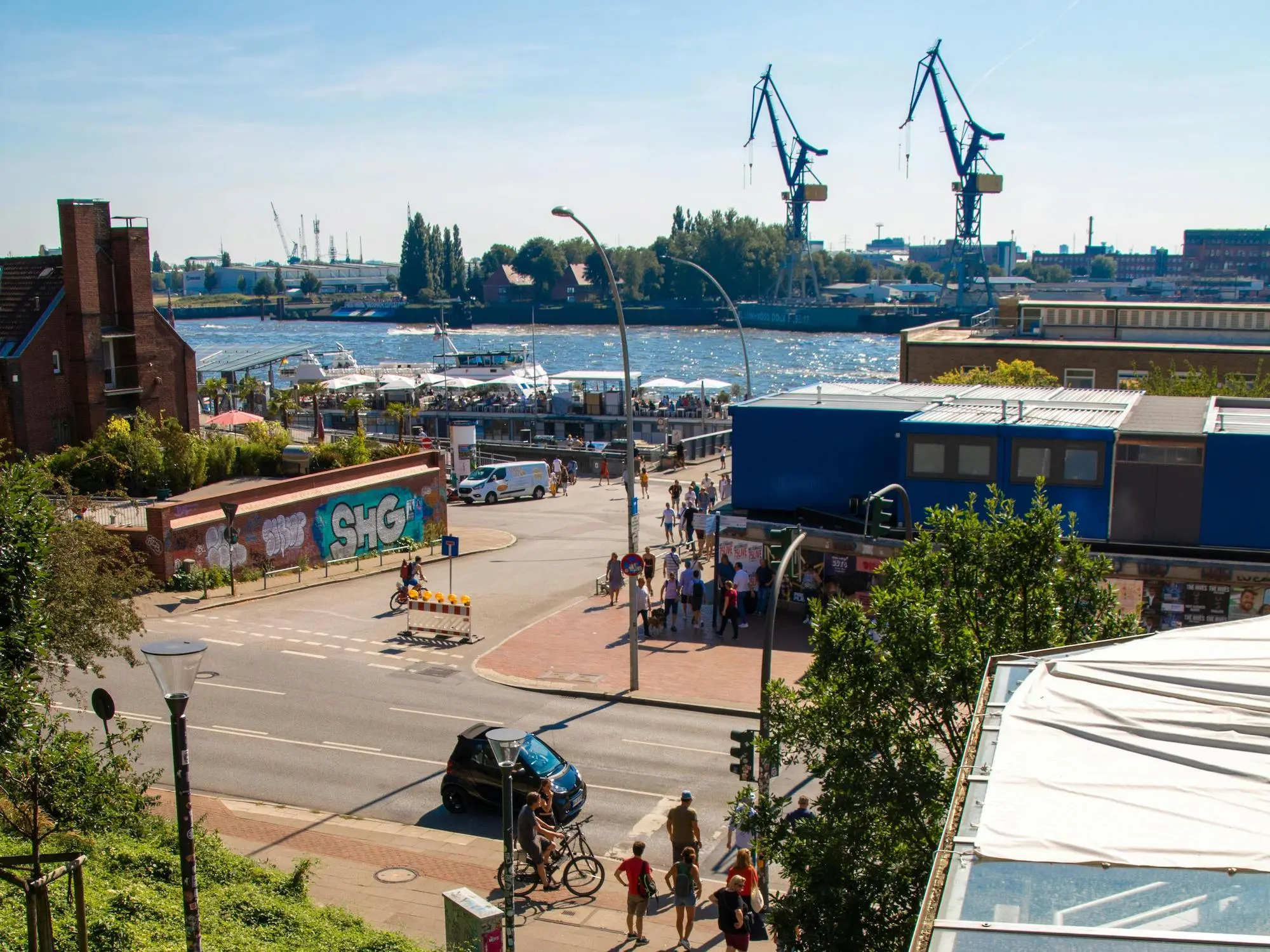 Menschen in Hamburg spazieren oder fahren mit Auto und Fahrrad durch die Straßen, im Hintergrund ist die Elbe und der Hafen zu sehen